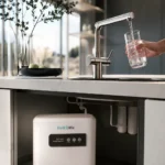 Person fills a glass from a modern kitchen faucet; a Hydrolife under-sink water filtration unit is visible beneath the counter.